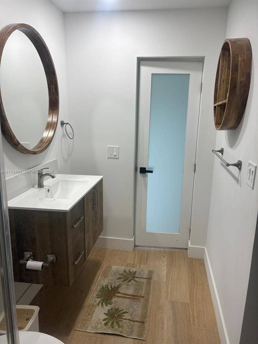 9961 SW 20th Street, Miami, Miami, FL 33165 Floating wood-finish vanity with an integrated basin, round wood-frame mirror, wood-finish wall shelving, wood-finish flooring, and a single-panel door with a frosted glass insert