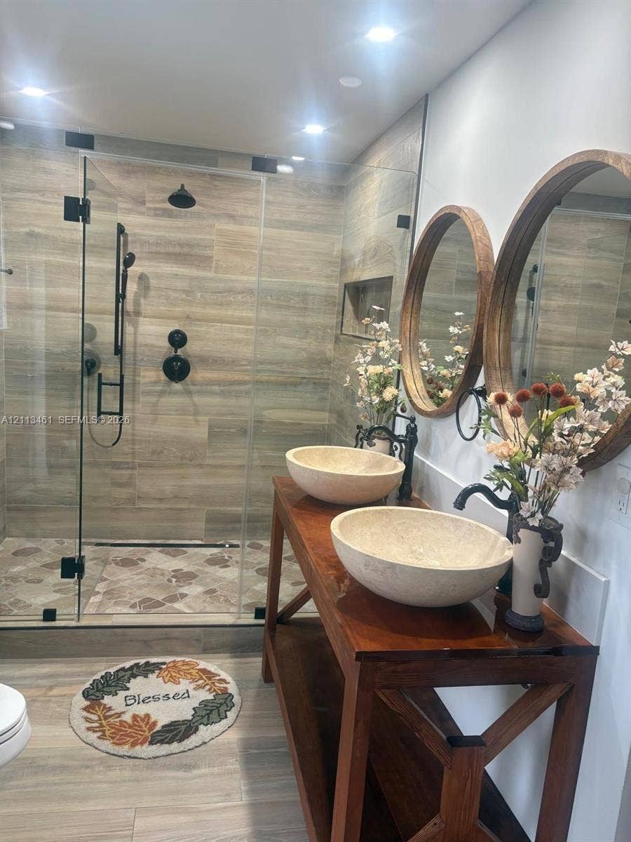 9961 SW 20th Street, Miami, Miami, FL 33165 Double vanity featuring natural stone vessel sinks, wooden base cabinetry, and circular framed mirrors, paired with a glass-enclosed shower featuring stone-look tile walls and matte black fixtures