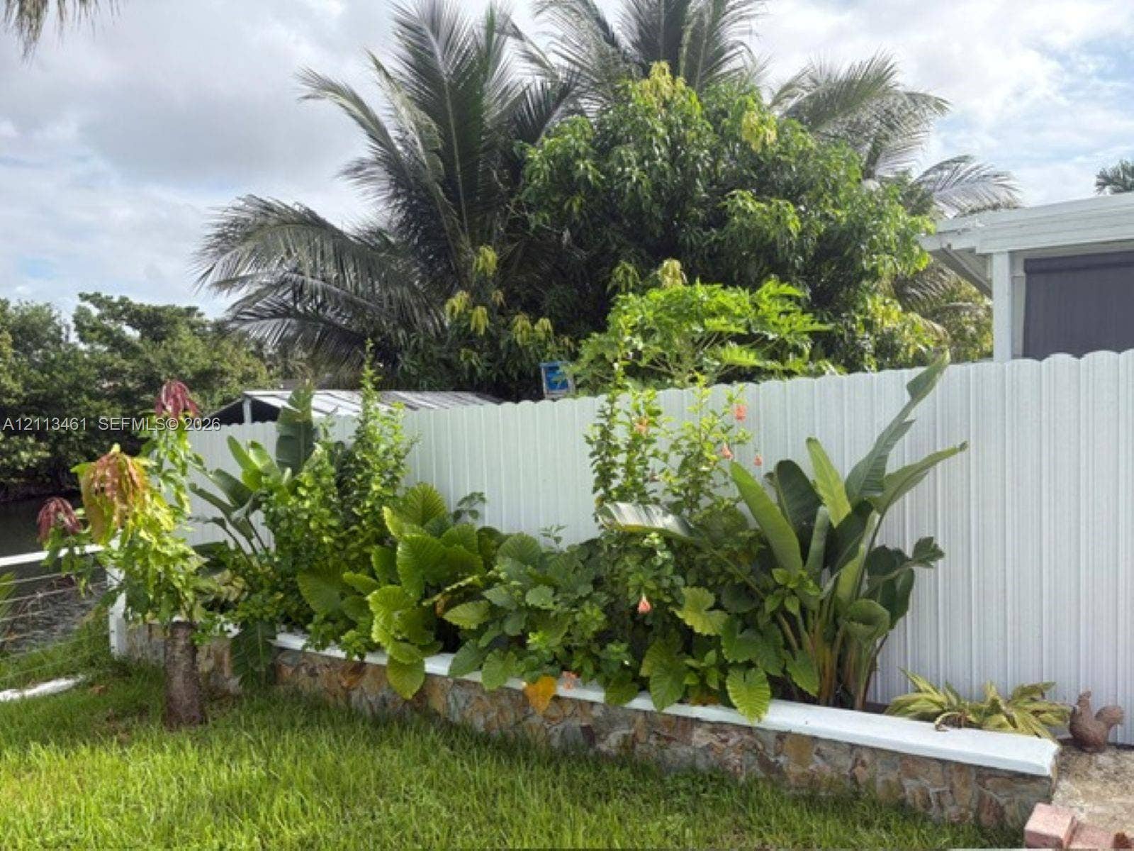 9961 SW 20th Street, Miami, Miami, FL 33165 Raised stone-veneer garden bed with a white cap, accented by mature tropical landscaping and a white vinyl privacy fence