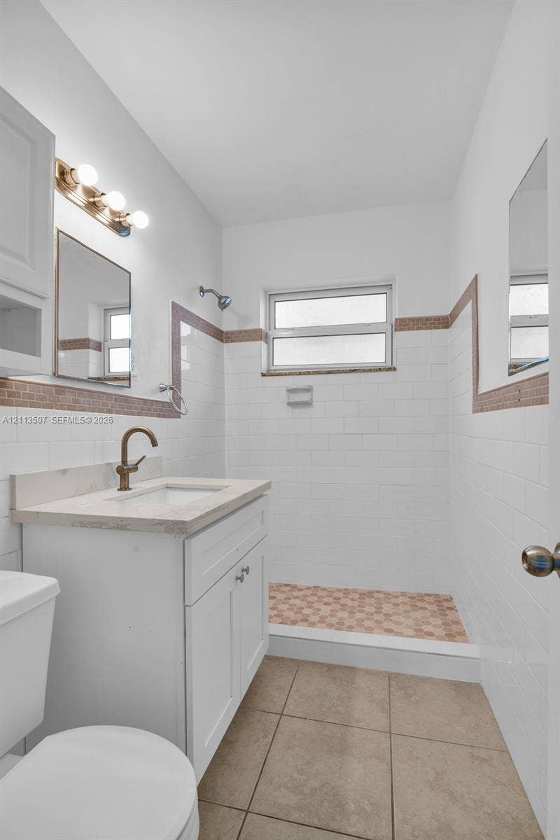 Bathroom featuring a white vanity with a neutral stone countertop, bronze-finish faucet, white subway tile walls with decorative border accents, walk-in shower with mosaic tile flooring, and large-format tile flooring