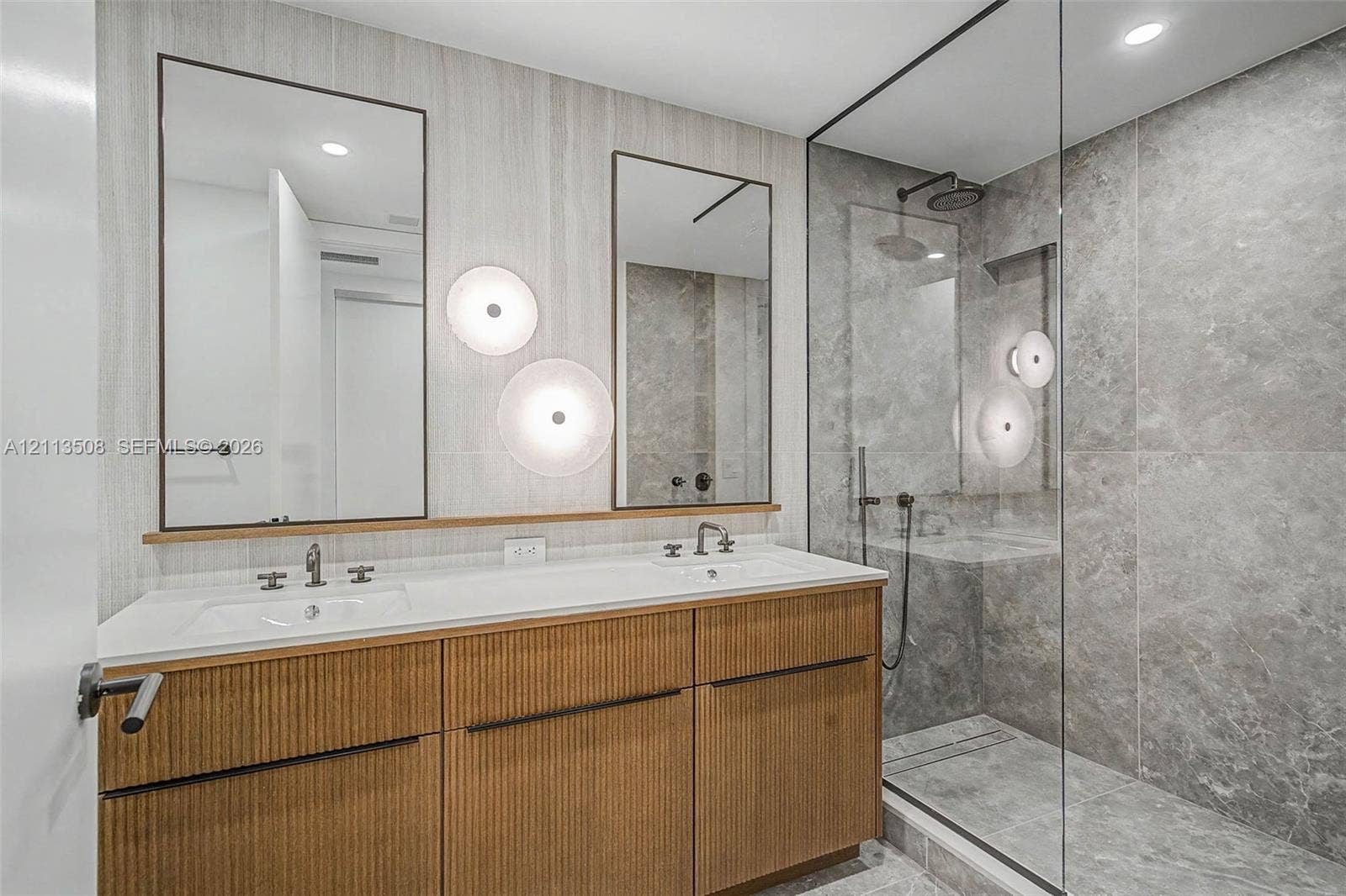 2200 Brickell Avenue, #405, Miami, Miami, FL 33129 Dual-sink vanity with fluted wood-finish cabinetry, rectangular under-mount basins, wall-mounted round lighting, and a glass-enclosed shower featuring large-format stone-style tile and rain-style showerhead