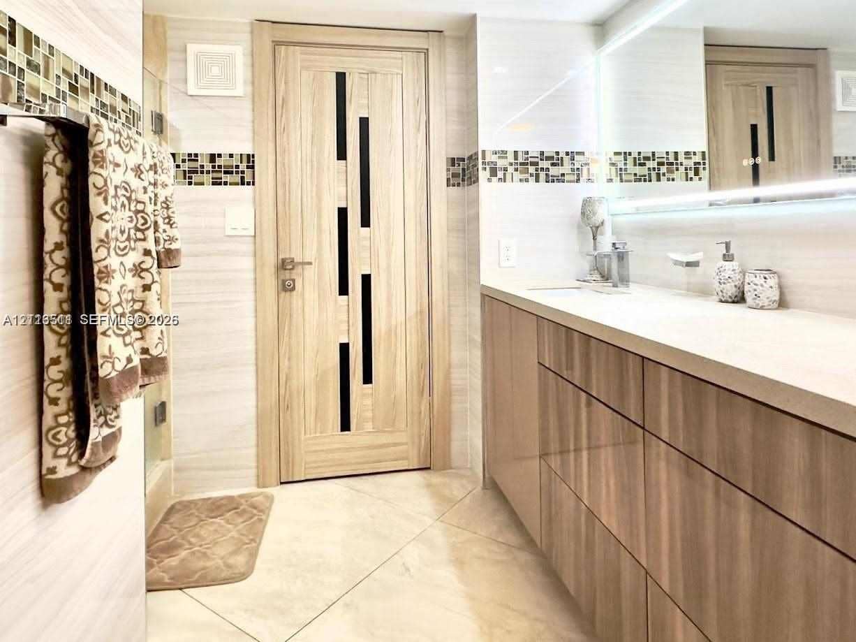 1201 S Ocean Drive, #2202S, Hollywood, FL 33019 Bathroom featuring a floating vanity with wood-finish cabinetry, a large wall-to-wall mirror with integrated lighting, glass-tile decorative inlay, and a modern interior door with vertical dark-finish glass accents