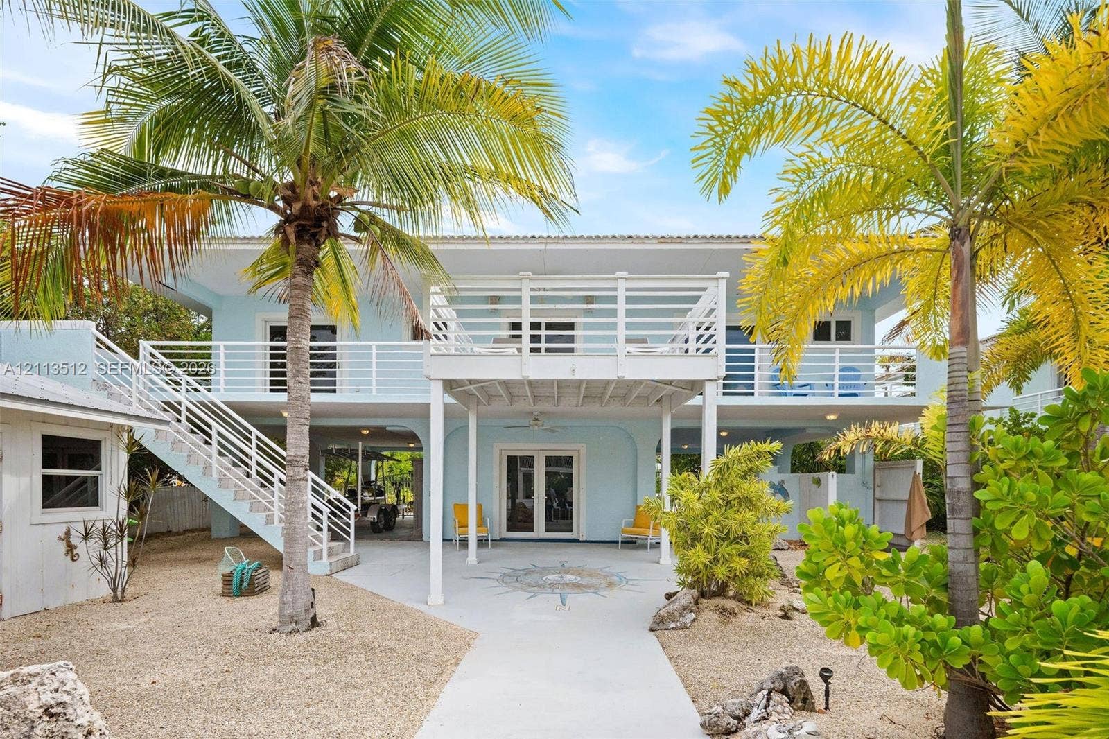 Key Largo, FL 33037 Elevated two-story residence featuring a ground-level covered patio with a decorative stone compass medallion, an expansive second-story balcony with white horizontal railings, an external staircase, and mature palm landscaping