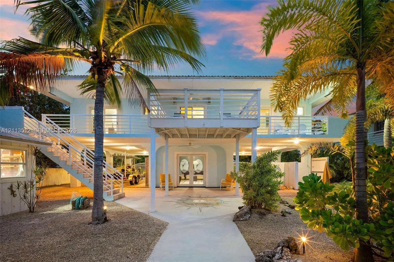 Key Largo, FL 33037 Two-story coastal residence featuring a stilt-construction design, a full-length elevated balcony with horizontal railings, an exterior staircase, and a ground-level covered patio with decorative stonework