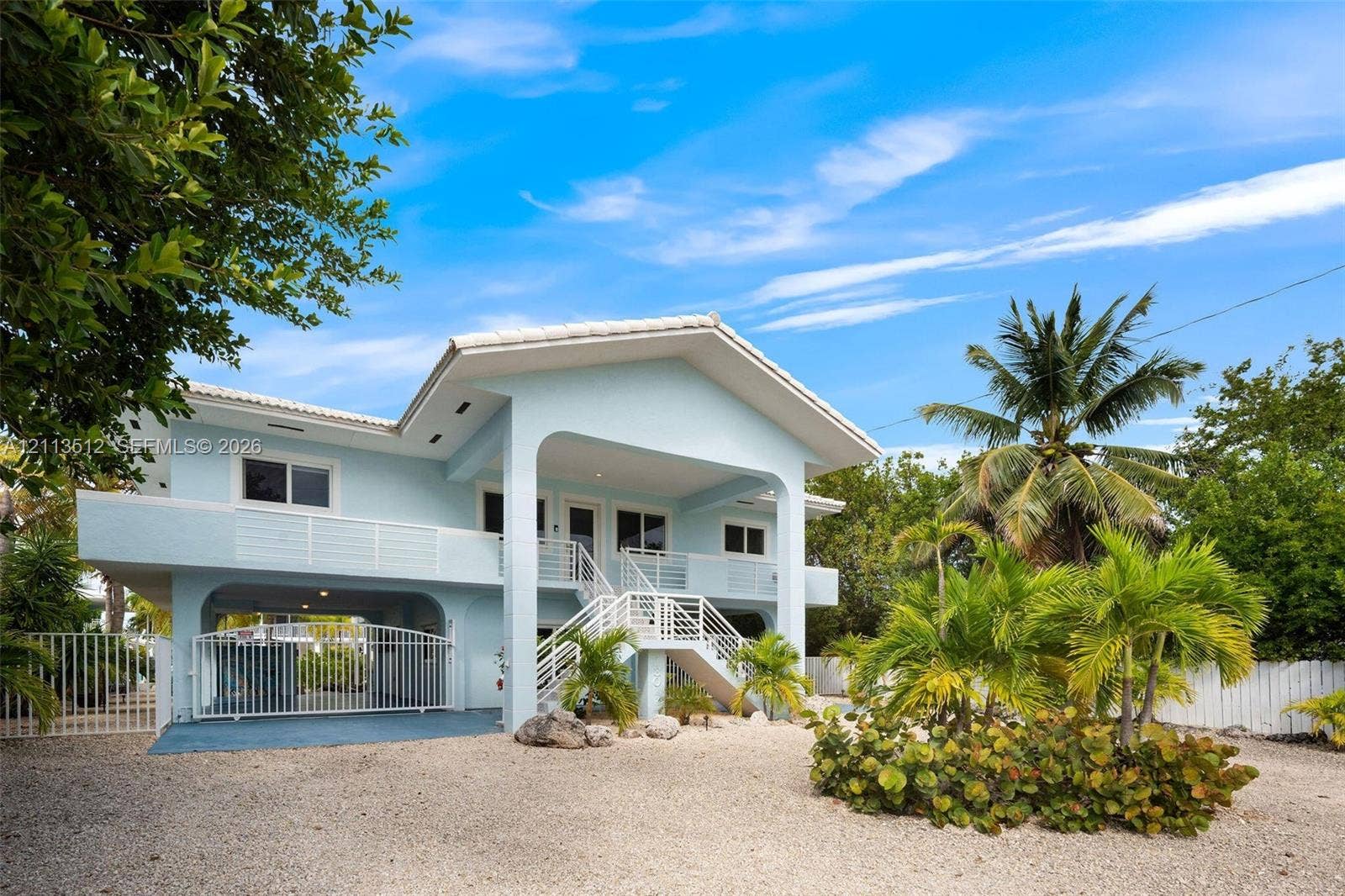 Key Largo, FL 33037 Elevated residence featuring a covered front porch, gated ground-level parking, metal railings, and a gravel driveway with mature landscaping