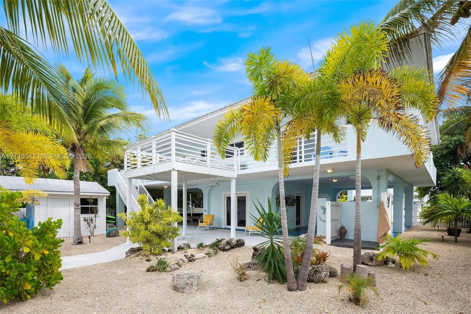 Key Largo, FL 33037 Elevated home exterior featuring a raised wooden balcony, covered ground-level patio, light-toned siding, and surrounding xeriscaped grounds with mature palm trees