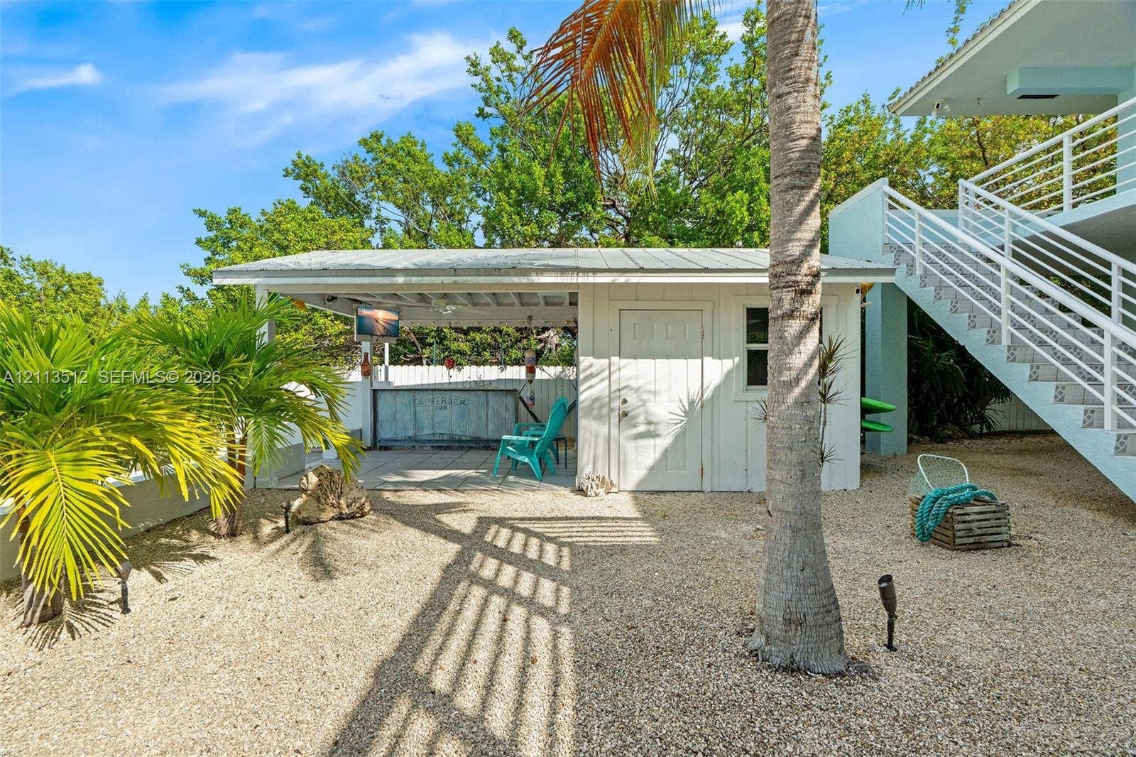 Key Largo, FL 33037 Outdoor covered bar area featuring a metal roof and concrete structure, surrounded by gravel grounds with mature palm trees and exterior stairs with white railing