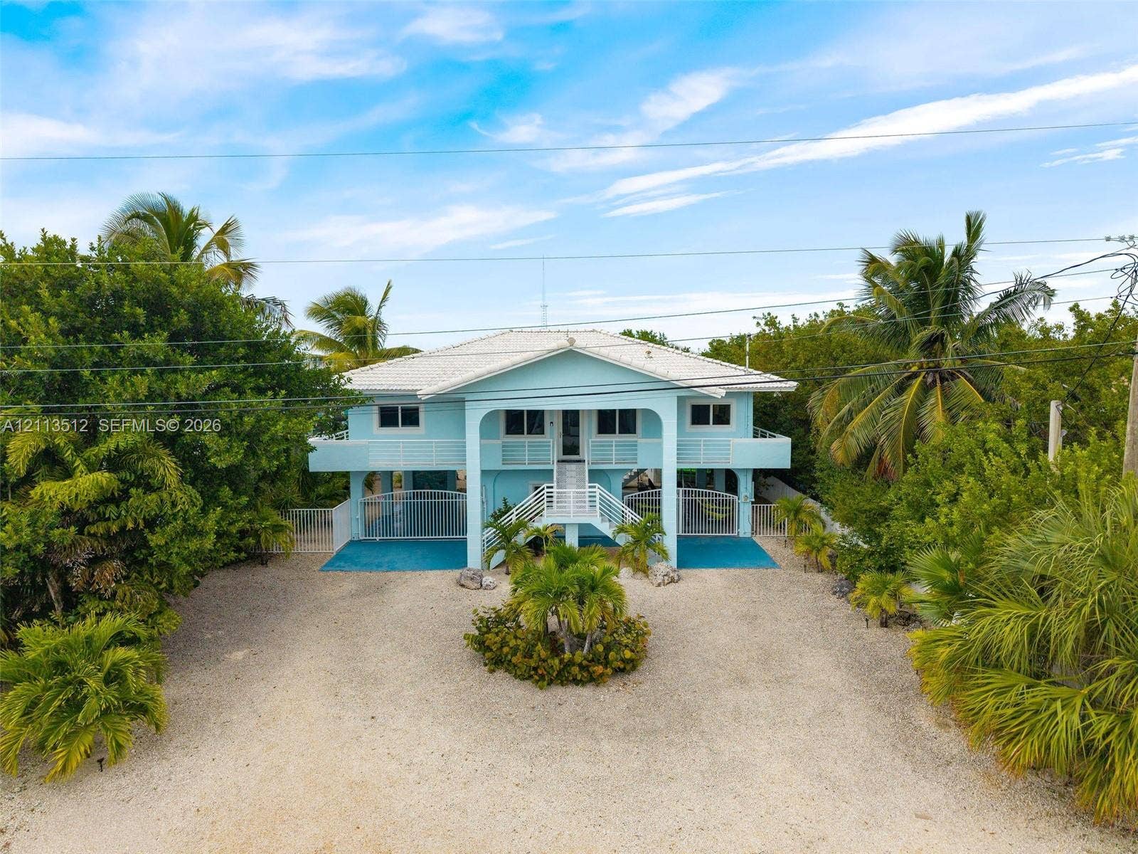 Key Largo, FL 33037 Elevated coastal residence featuring a metal roof, wraparound balcony, central staircase with white railing, and gravel driveway
