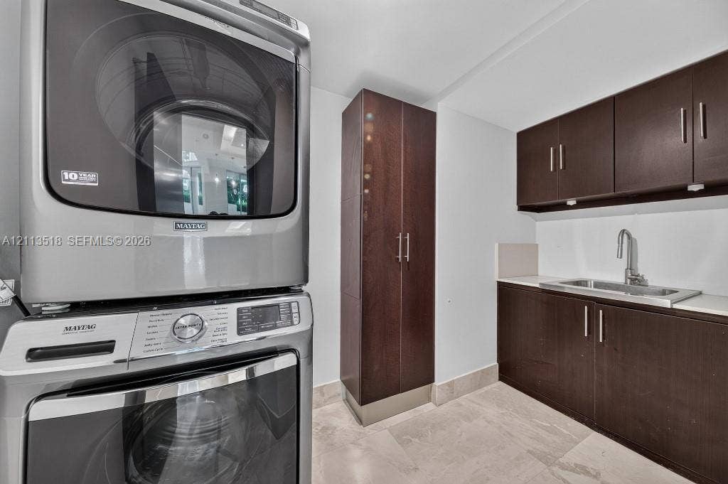 3131 NE 188th Street, #1-303, Aventura, FL 33180 Laundry area featuring dark wood-finish cabinetry, a tall pantry unit, a stainless steel undermount sink with a gooseneck faucet, light-toned stone-finish flooring, and a stacked front-loading washer and dryer set