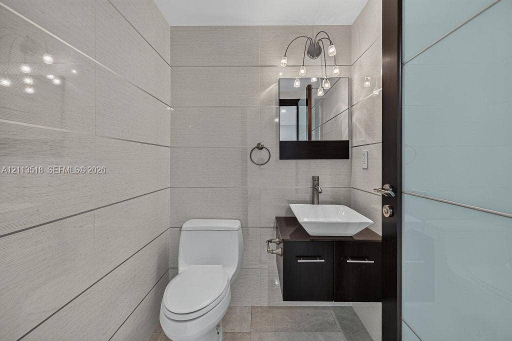 3131 NE 188th Street, #1-303, Aventura, FL 33180 Bathroom featuring floor-to-ceiling large-format tile, wall-mounted dark wood vanity with a white vessel sink, multi-bulb wall-mounted lighting, and a frosted glass panel door