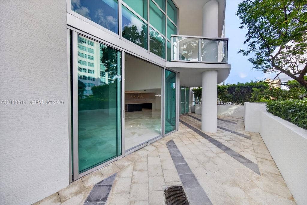 3131 NE 188th Street, #1-303, Aventura, FL 33180 Expansive stone-paved terrace with contrasting linear accents, floor-to-ceiling glass sliding doors, elevated glass-paneled balcony, and structural concrete columns