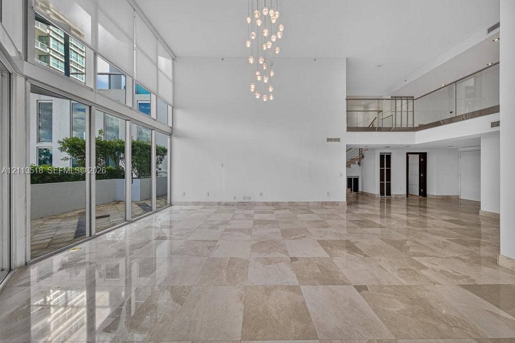 3131 NE 188th Street, #1-303, Aventura, FL 33180 Double-height ceilings, floor-to-ceiling glass wall, polished stone flooring, multi-tiered pendant light fixture, and second-level mezzanine with glass railing