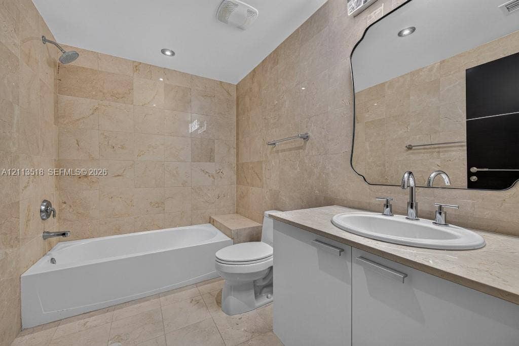 3131 NE 188th Street, #1-303, Aventura, FL 33180 Full-height natural stone wall tiling, white vanity with an integrated countertop, oval undermount sink, decorative wall mirror, and built-in tub surround