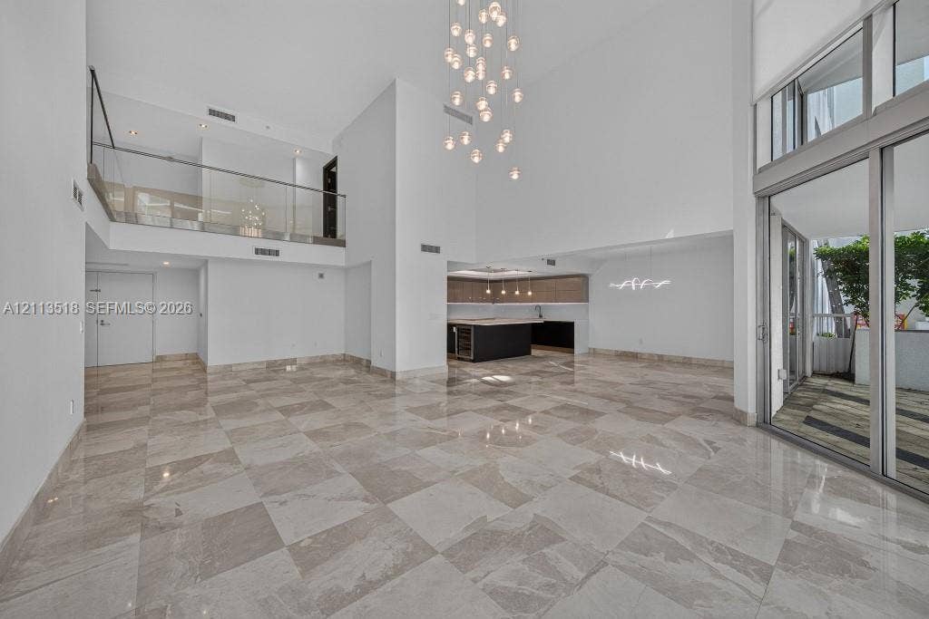 3131 NE 188th Street, #1-303, Aventura, FL 33180 Open-concept interior featuring double-height ceilings, stone-finish tile flooring, a glass-panel mezzanine railing, multi-light pendant chandelier, and expansive floor-to-ceiling sliding glass doors