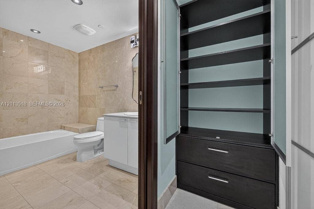 3131 NE 188th Street, #1-303, Aventura, FL 33180 Built-in cabinetry featuring dark wood-finish shelving and drawers with frosted glass doors, complemented by a bathroom finished with stone-tile wall and floor surfacing, a white vanity cabinet, and a built-in bathtub