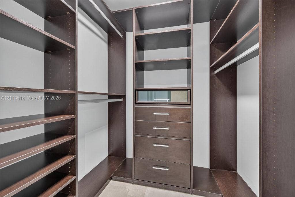 3131 NE 188th Street, #1-303, Aventura, FL 33180 Custom walk-in storage system featuring dark wood-finish cabinetry, built-in shelving, integrated hanging rods, and a centralized unit with four drawers and a glass-front display compartment