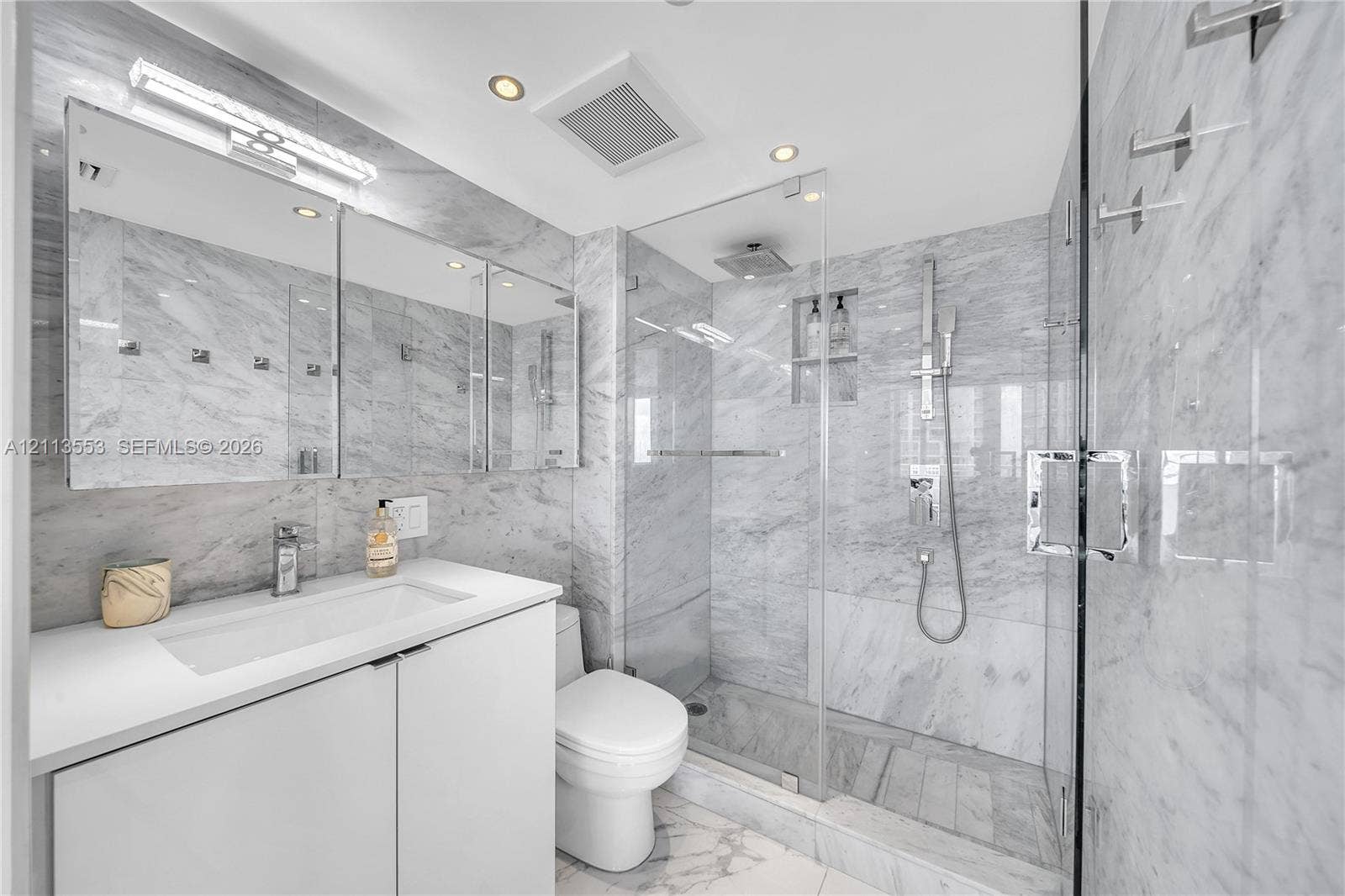 400 S Pointe Drive, #2106, Miami Beach, FL 33139 Bathroom featuring wall-to-wall natural stone tiling, a glass-enclosed walk-in shower with integrated shelving, a modern vanity with a white countertop, and a recessed medicine cabinet with integrated lighting