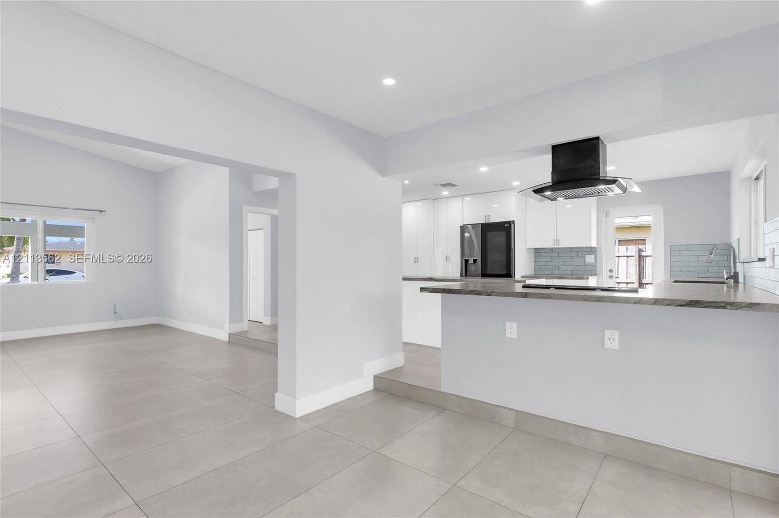 Open-concept kitchen featuring a breakfast bar, white cabinetry, dark stone-finish countertops, tile backsplash, and a black range hood