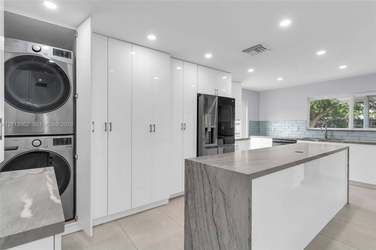 Kitchen featuring a waterfall-edge island with stone-finish countertop, high-gloss white cabinetry, integrated laundry station, and recessed lighting