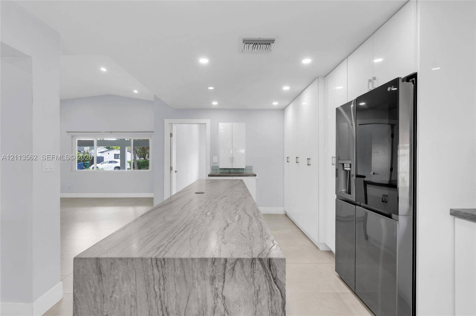 Kitchen featuring a large central island with waterfall-edge countertop, floor-to-ceiling high-gloss white cabinetry, large-format tile flooring, and integrated stainless steel refrigerator