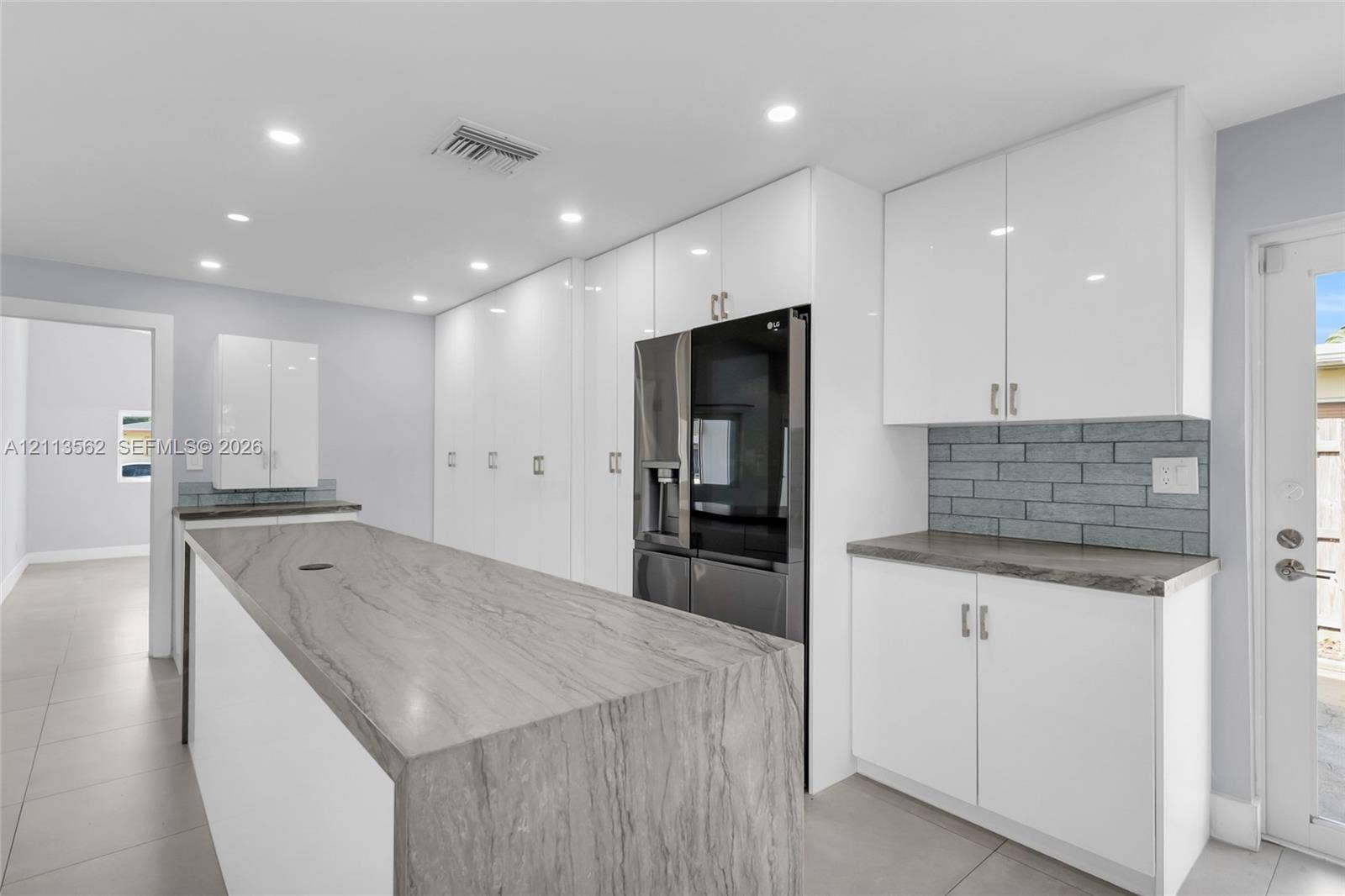 Kitchen featuring white flat-panel cabinetry, stone-surface waterfall island, grey brick-pattern backsplash, and tile flooring