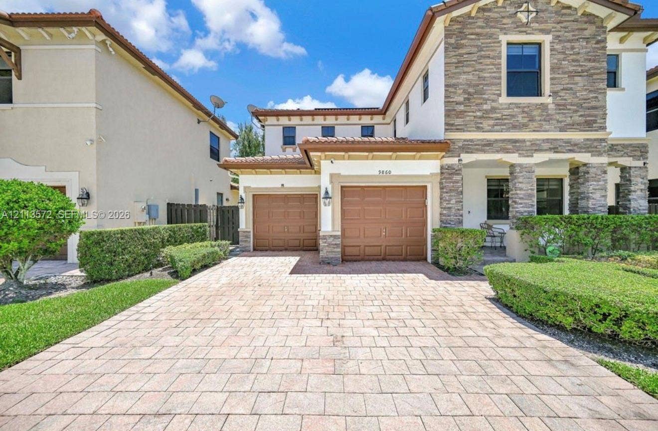9860 NW 86th Terrace, Doral, FL 33178 Two-story residence featuring a stone-veneer facade, paver driveway, three-car garage, tile roof, and manicured landscaping