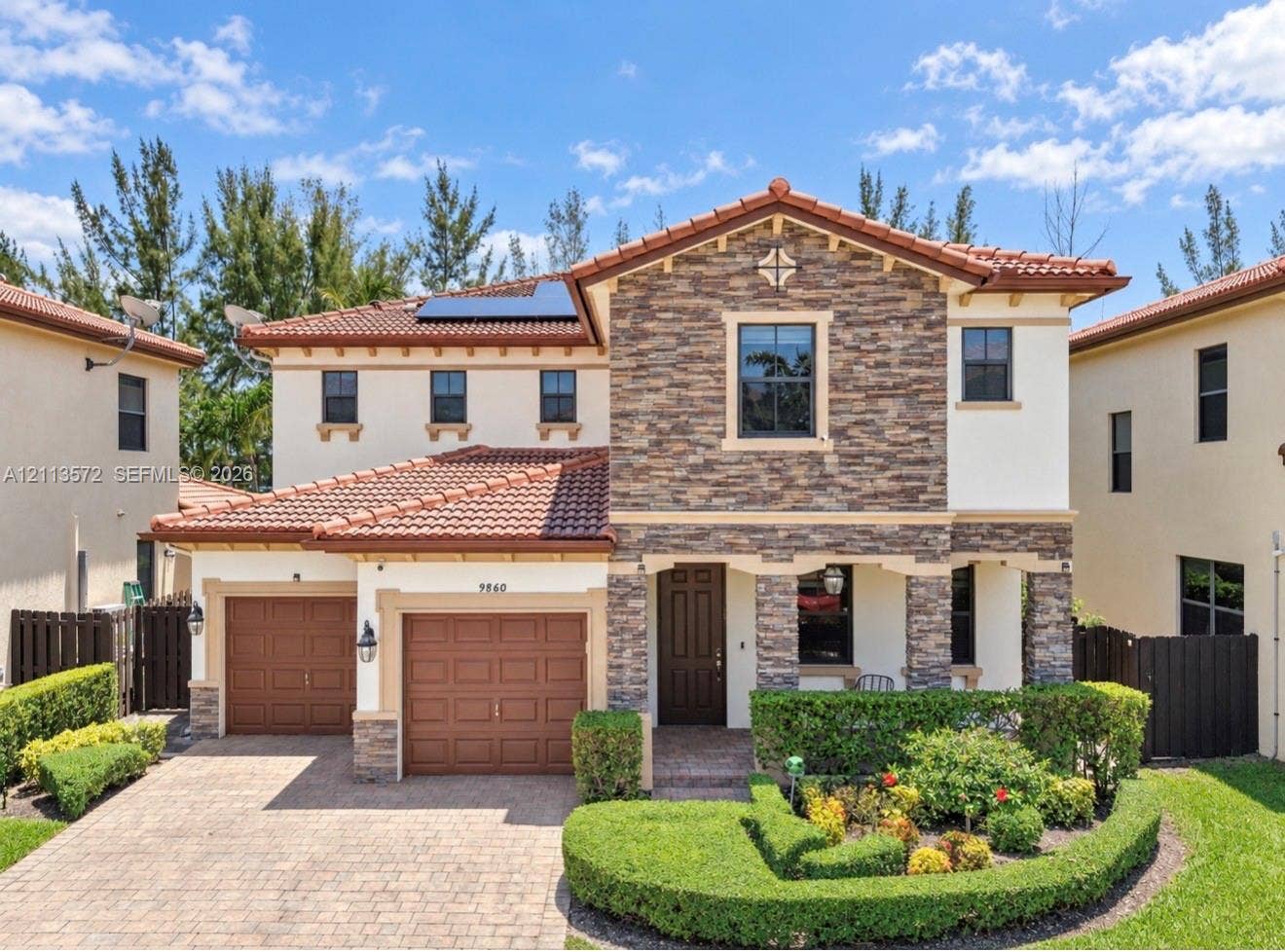 9860 NW 86th Terrace, Doral, FL 33178 Two-story residence with a stone veneer facade, clay tile roof, dual-bay garage, solar panels, and a paver driveway