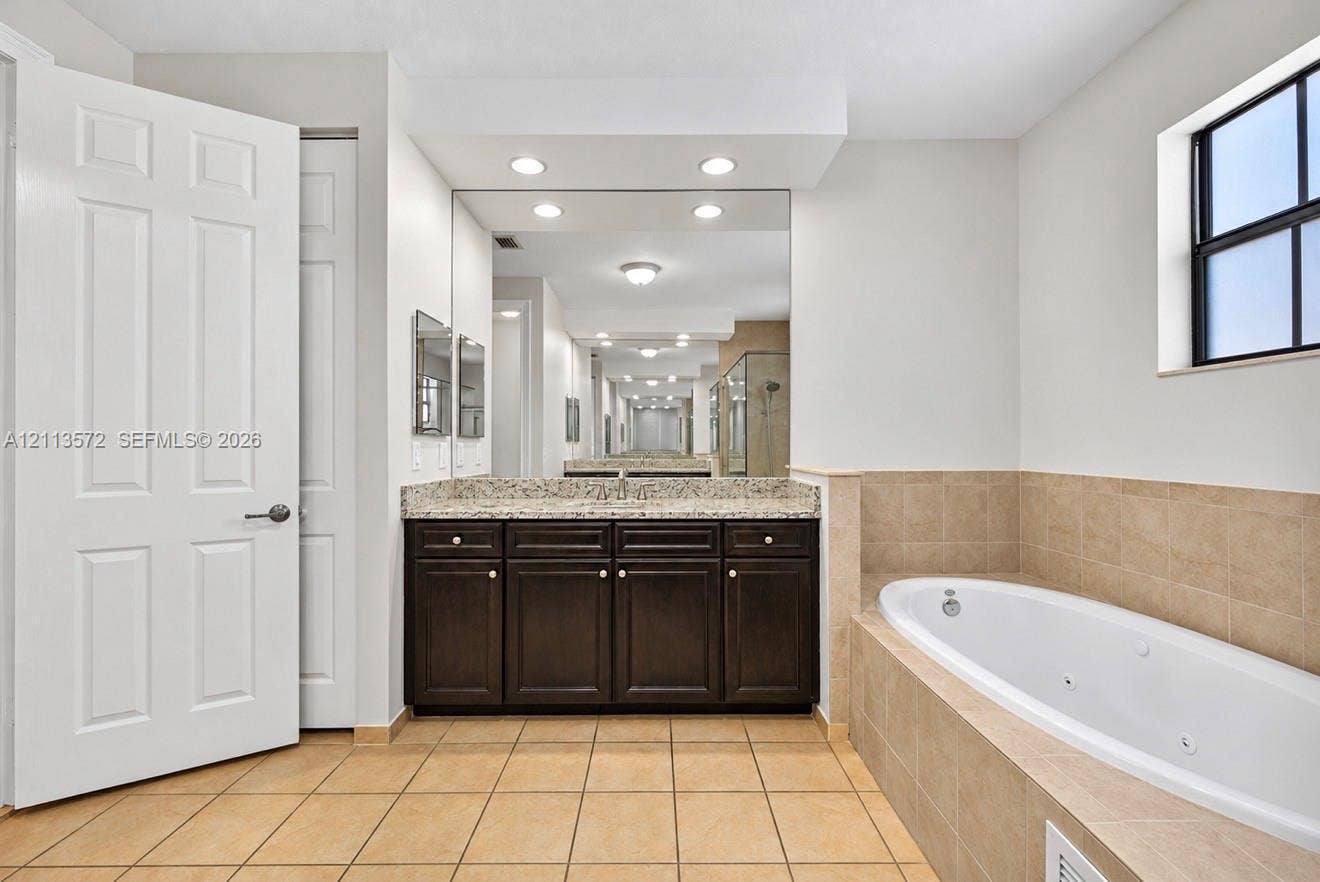 9860 NW 86th Terrace, Doral, FL 33178 Bathroom featuring a dark-finish vanity, stone-pattern countertop, recessed lighting, wall-to-wall mirror, and a jetted soaking tub with tile surround