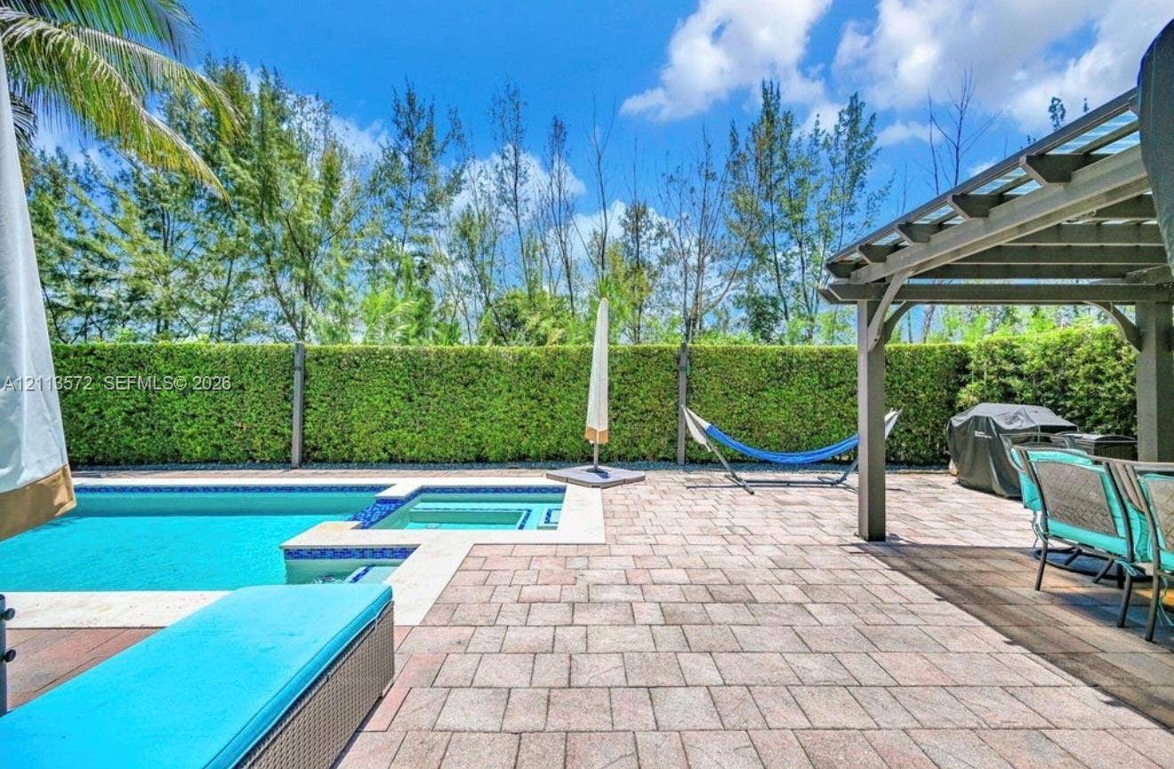 9860 NW 86th Terrace, Doral, FL 33178 Outdoor patio area featuring a paver stone deck, a covered pergola, an in-ground swimming pool with an integrated spa, and manicured hedge fencing