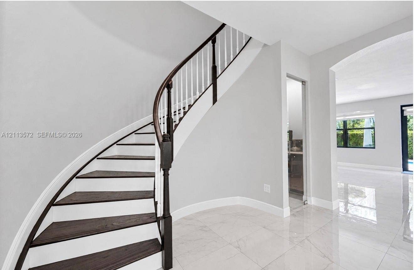 9860 NW 86th Terrace, Doral, FL 33178 Curved staircase with dark-finished treads and white risers, decorative wood handrail, polished stone flooring, and arched entryway
