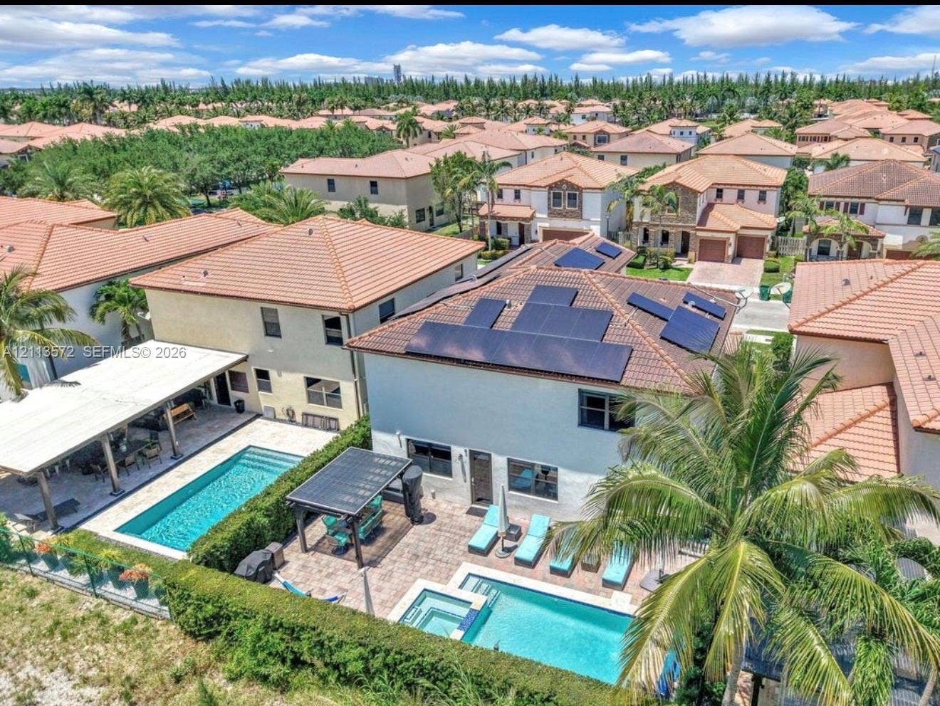 9860 NW 86th Terrace, Doral, FL 33178 Aerial perspective featuring a two-story residence equipped with roof-mounted solar panels, a rectangular swimming pool, a spillover spa, a stone-paved patio, and a covered lanai