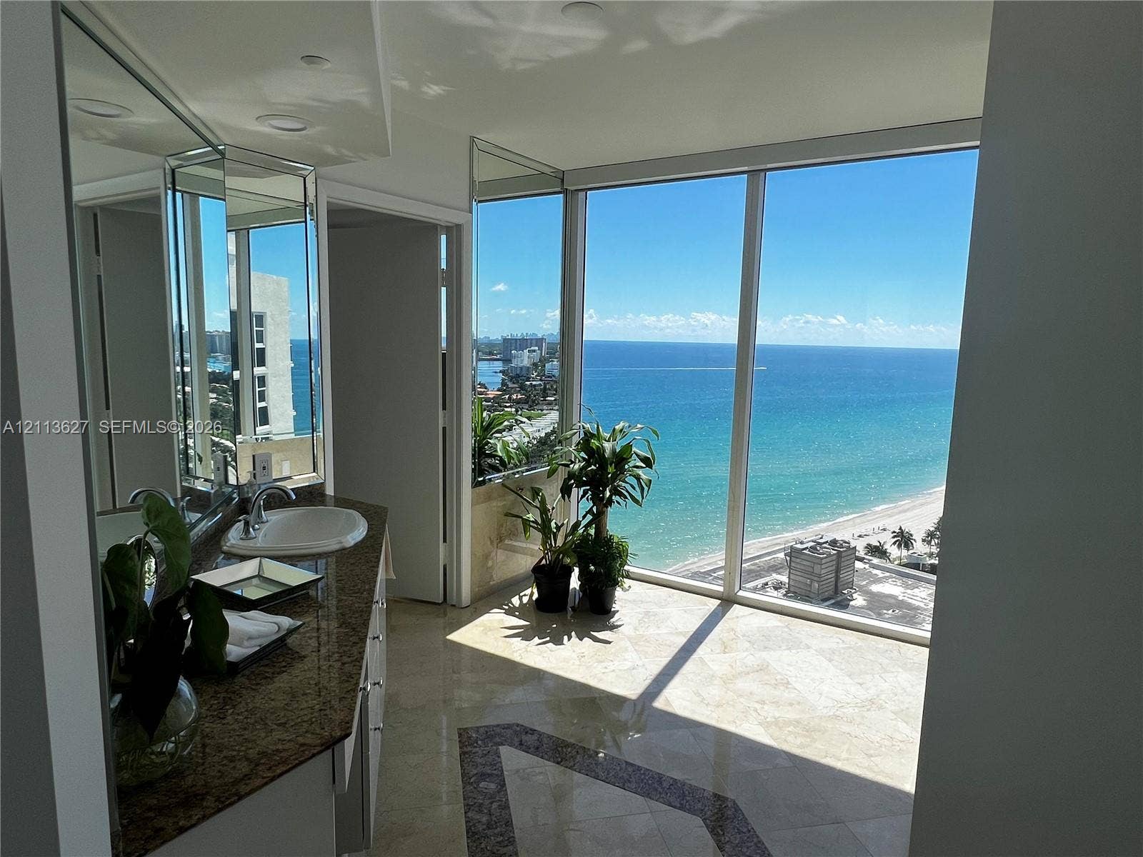 Floor-to-ceiling windows, stone tile flooring with decorative inlay, expansive vanity with integrated sink, and full-wall mirrors overlooking the ocean coastline