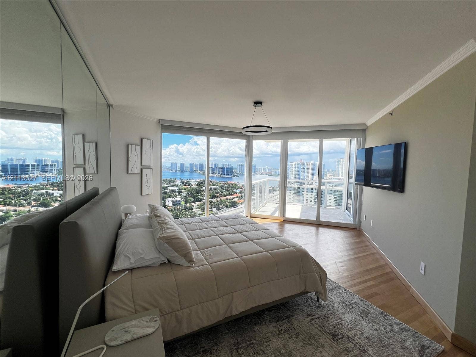 Floor-to-ceiling windows, wood-finish flooring, mirrored accent wall, circular pendant lighting, and an attached balcony with water and skyline scenery