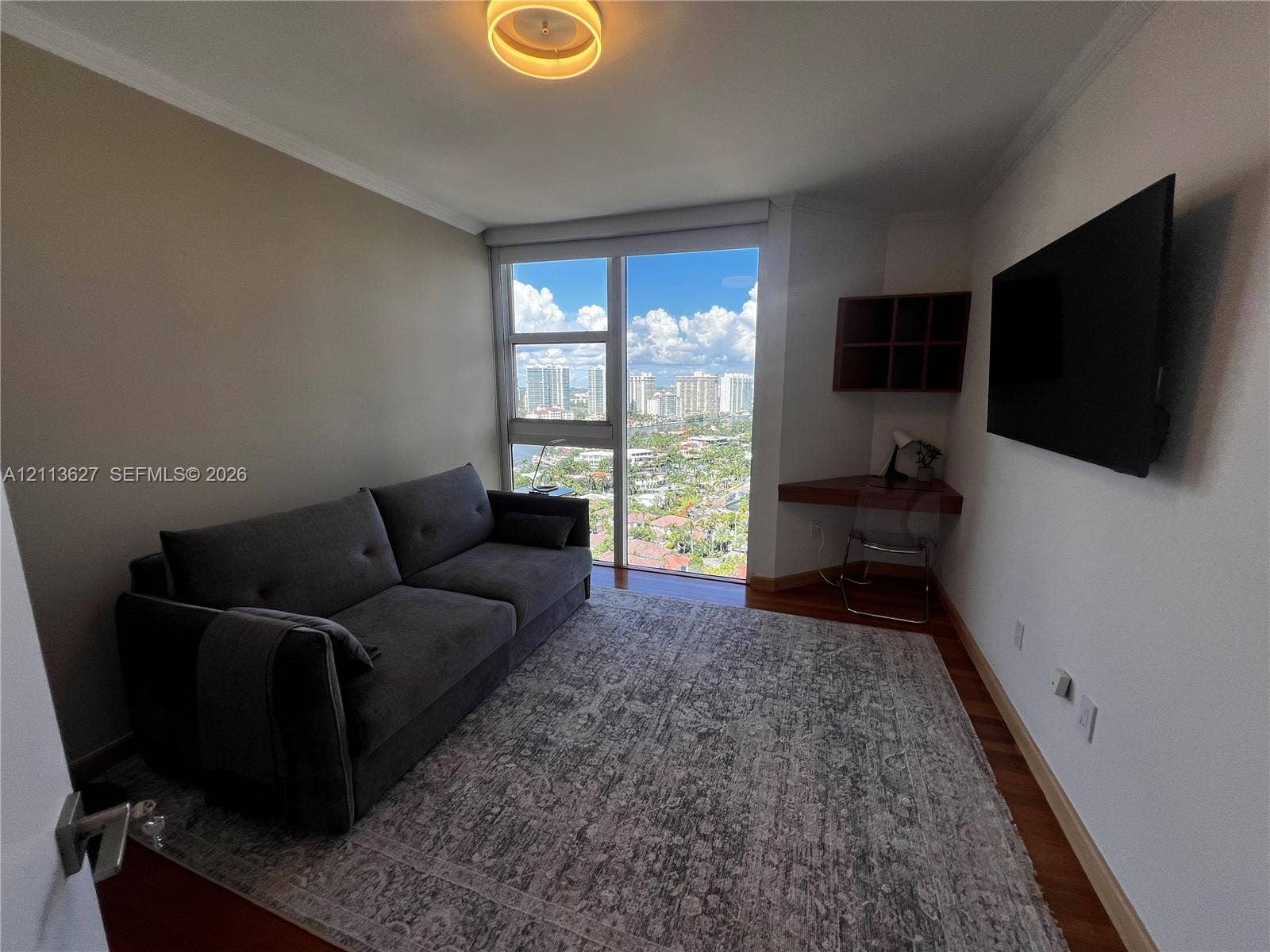 Floor-to-ceiling windows featuring urban scenery, built-in corner desk with integrated shelving, wood-finish flooring, and circular flush-mount ceiling lighting