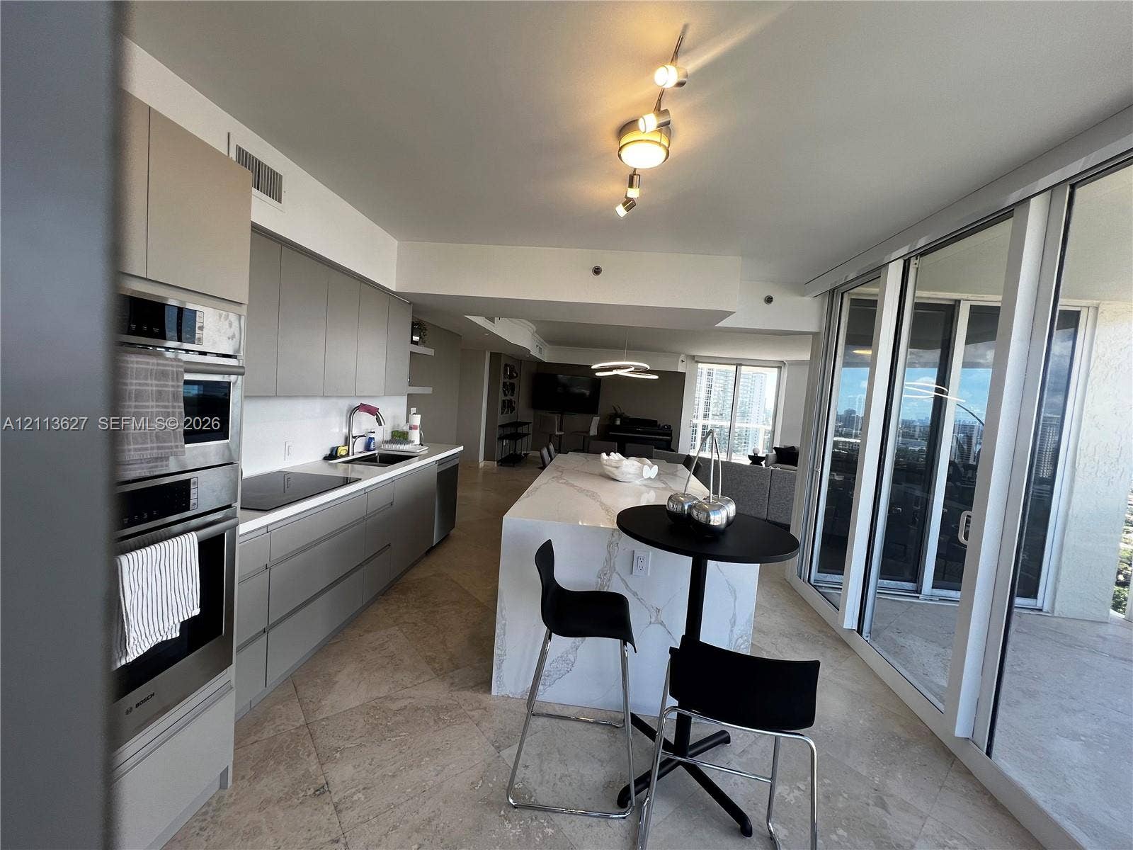 Contemporary kitchen featuring flat-panel cabinetry, a waterfall-edge stone island, built-in stainless steel appliances, stone flooring, and expansive sliding glass doors