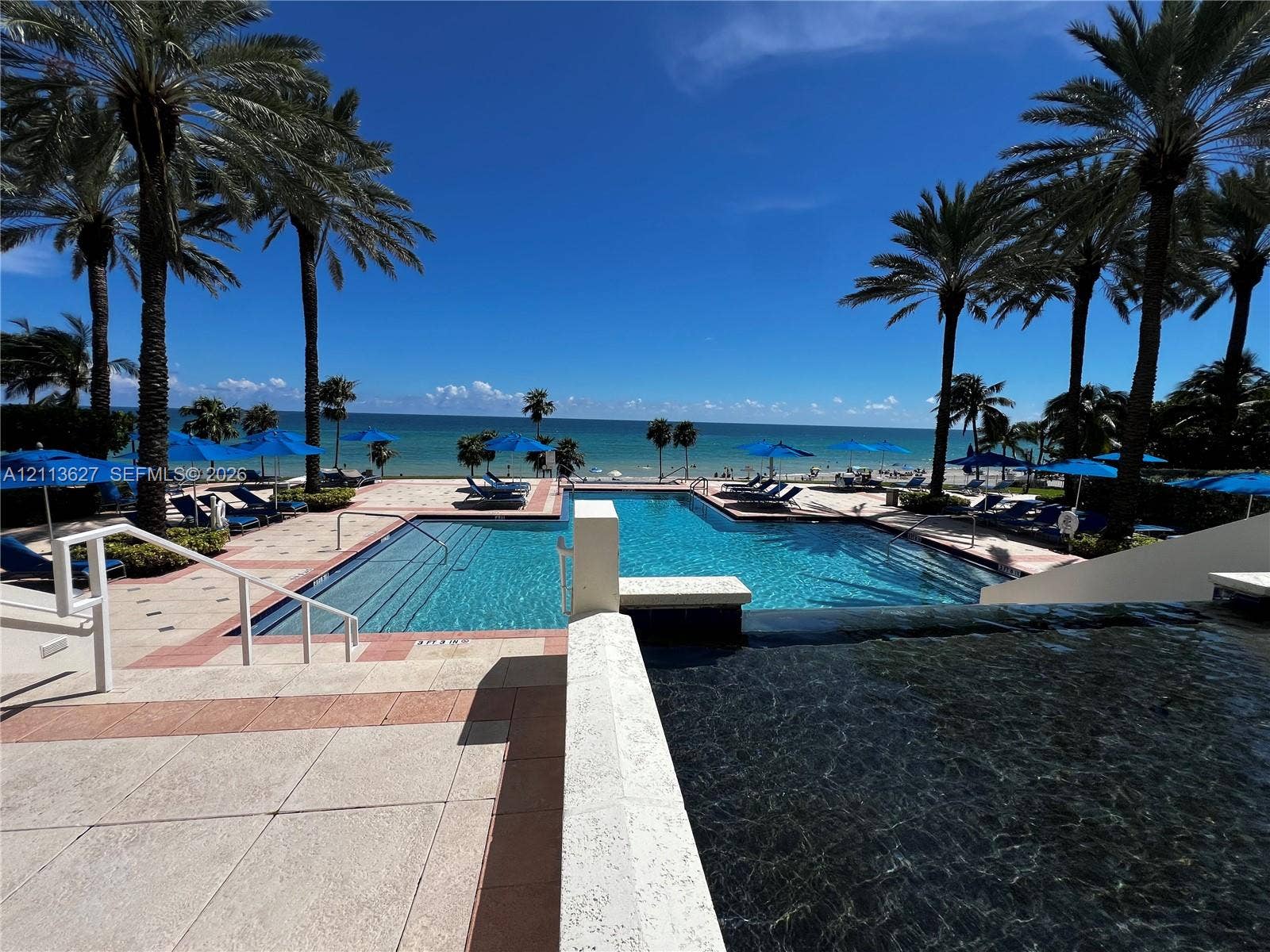 Outdoor pool area featuring a multi-level swimming pool, expansive stone-paver decking, integrated stairs with white metal handrails, and surrounding mature palm trees