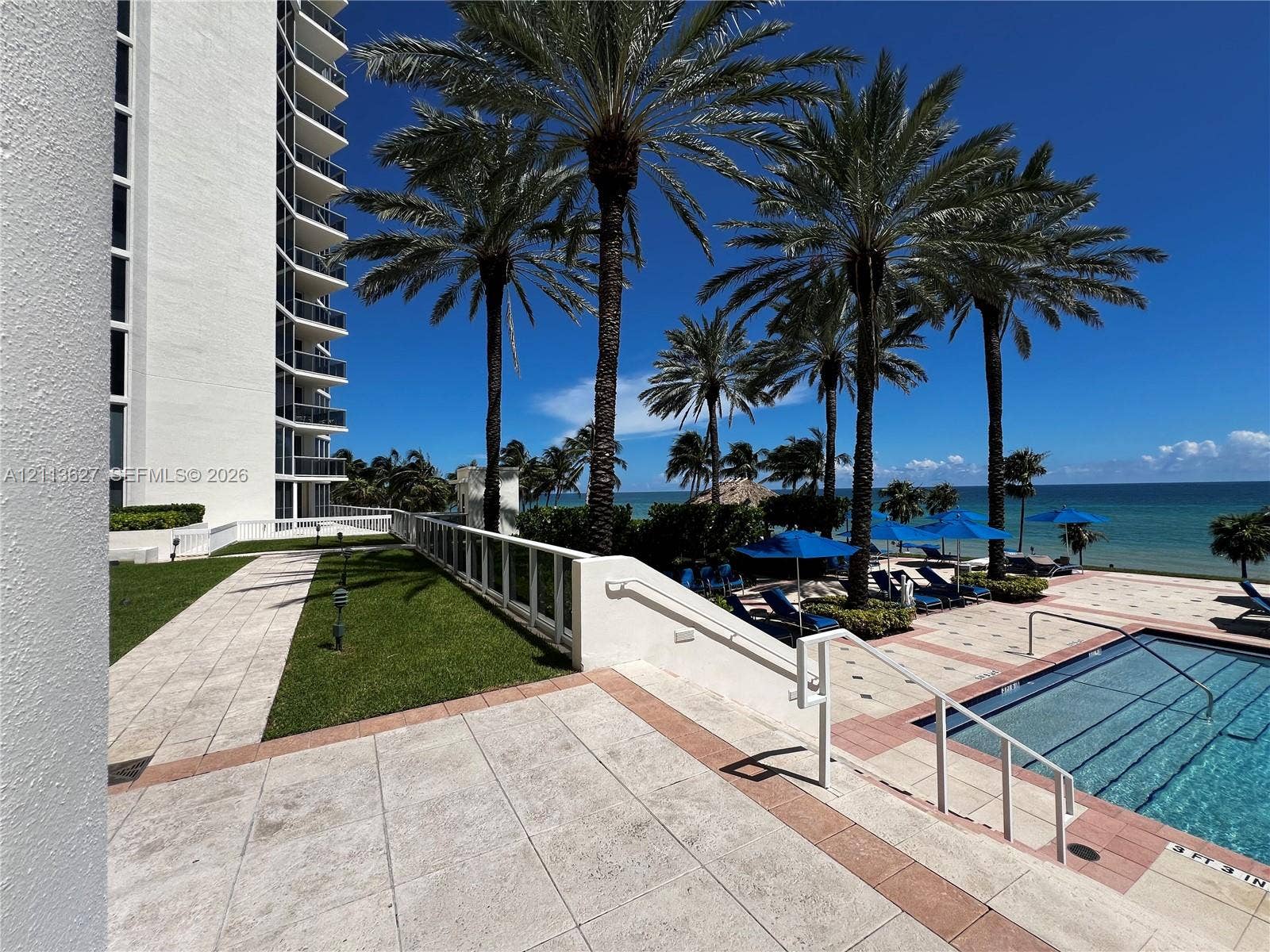 Outdoor swimming pool with tile decking, manicured lawn, palm trees, and barrier railing