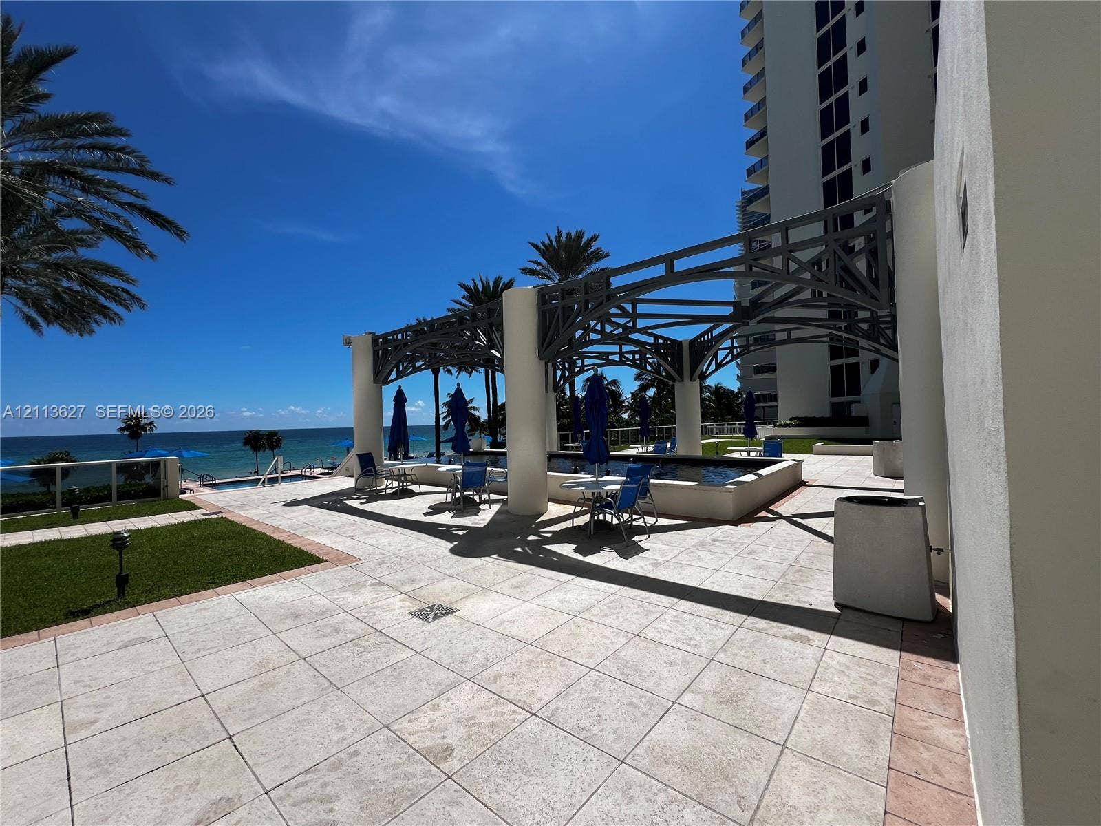 Outdoor terrace featuring stone tile flooring, a structured metal pergola, swimming pool, manicured lawn, and ocean backdrop