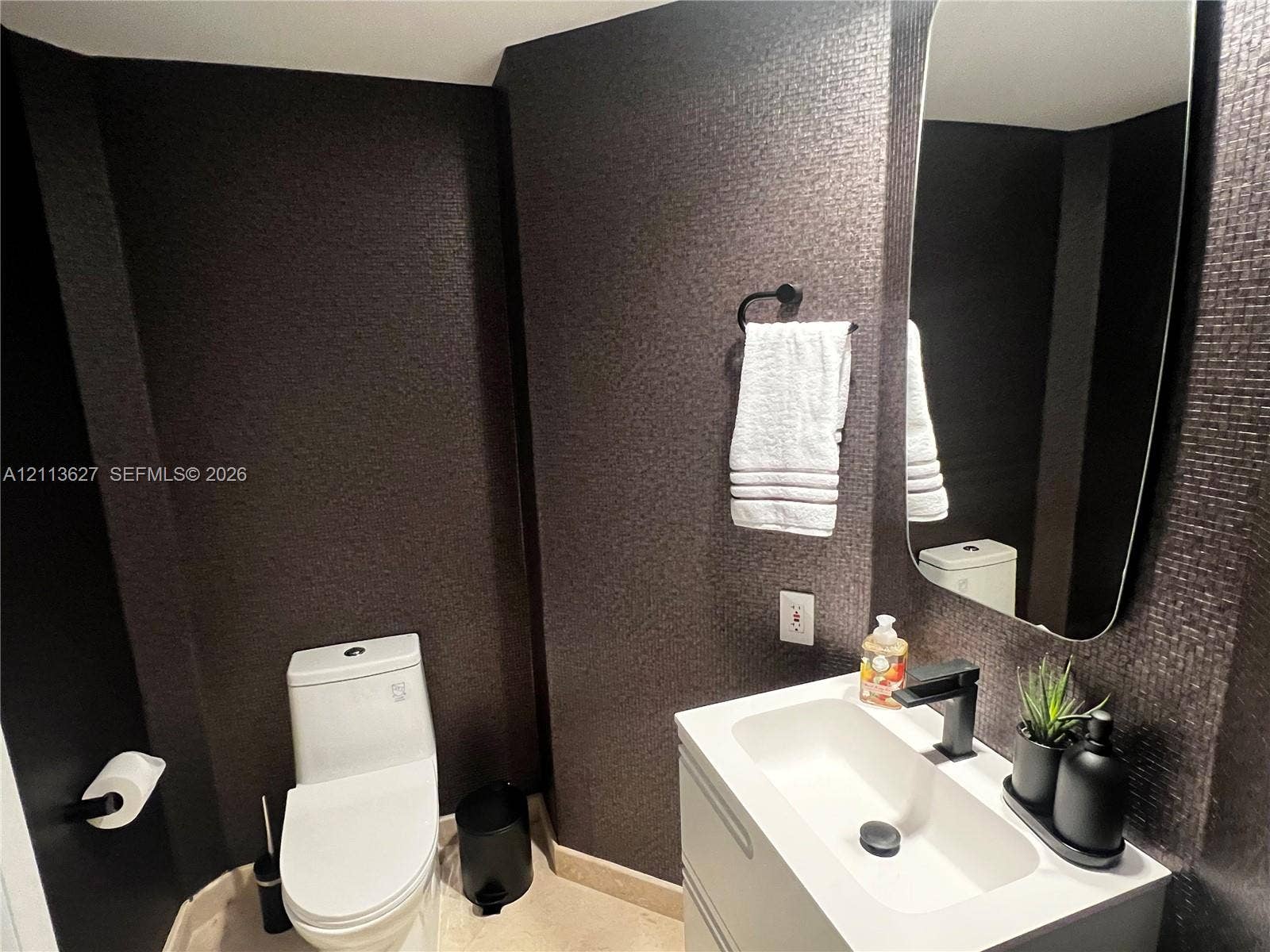 Modern floating vanity with a white integrated basin, matte black widespread faucet, textured wall covering, and a vertical rectangular mirror