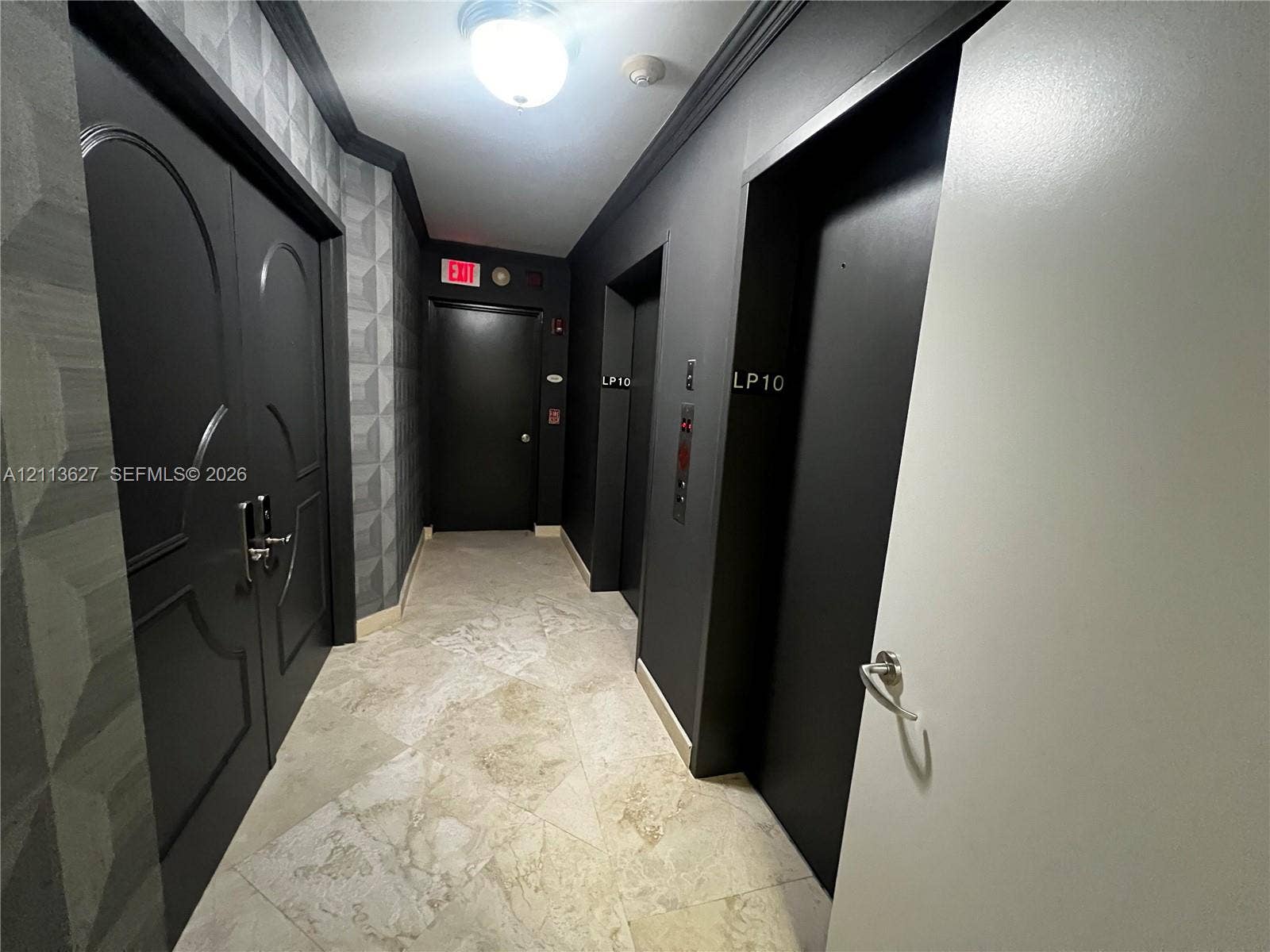 Stone tile flooring, geometric textured wall covering, double-door entry with metallic hardware, dual elevator access, and monochromatic architectural detailing
