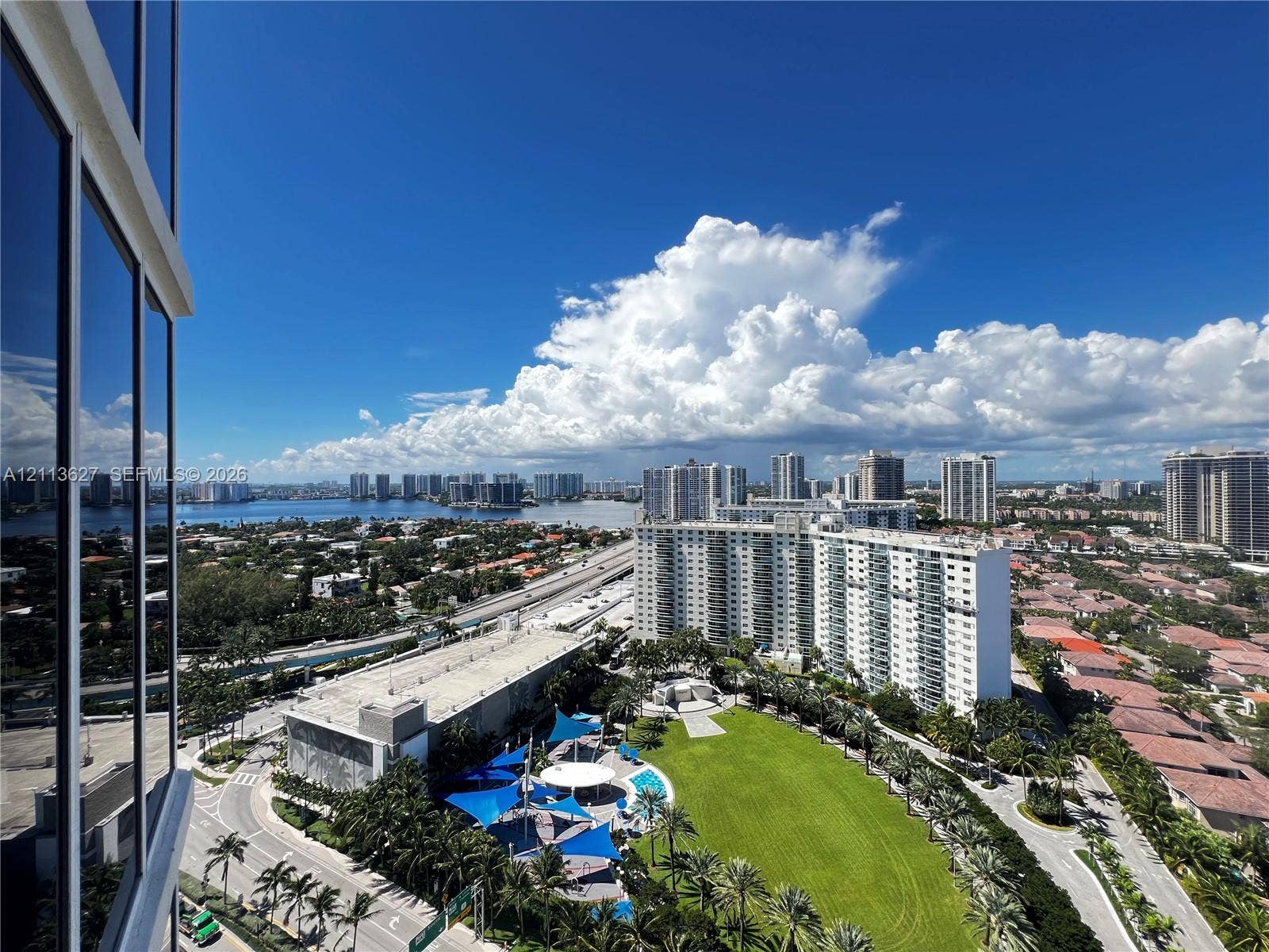 High-rise perspective featuring glass curtain wall facade, expansive green space with mature palms, and structured recreation area with shade sails