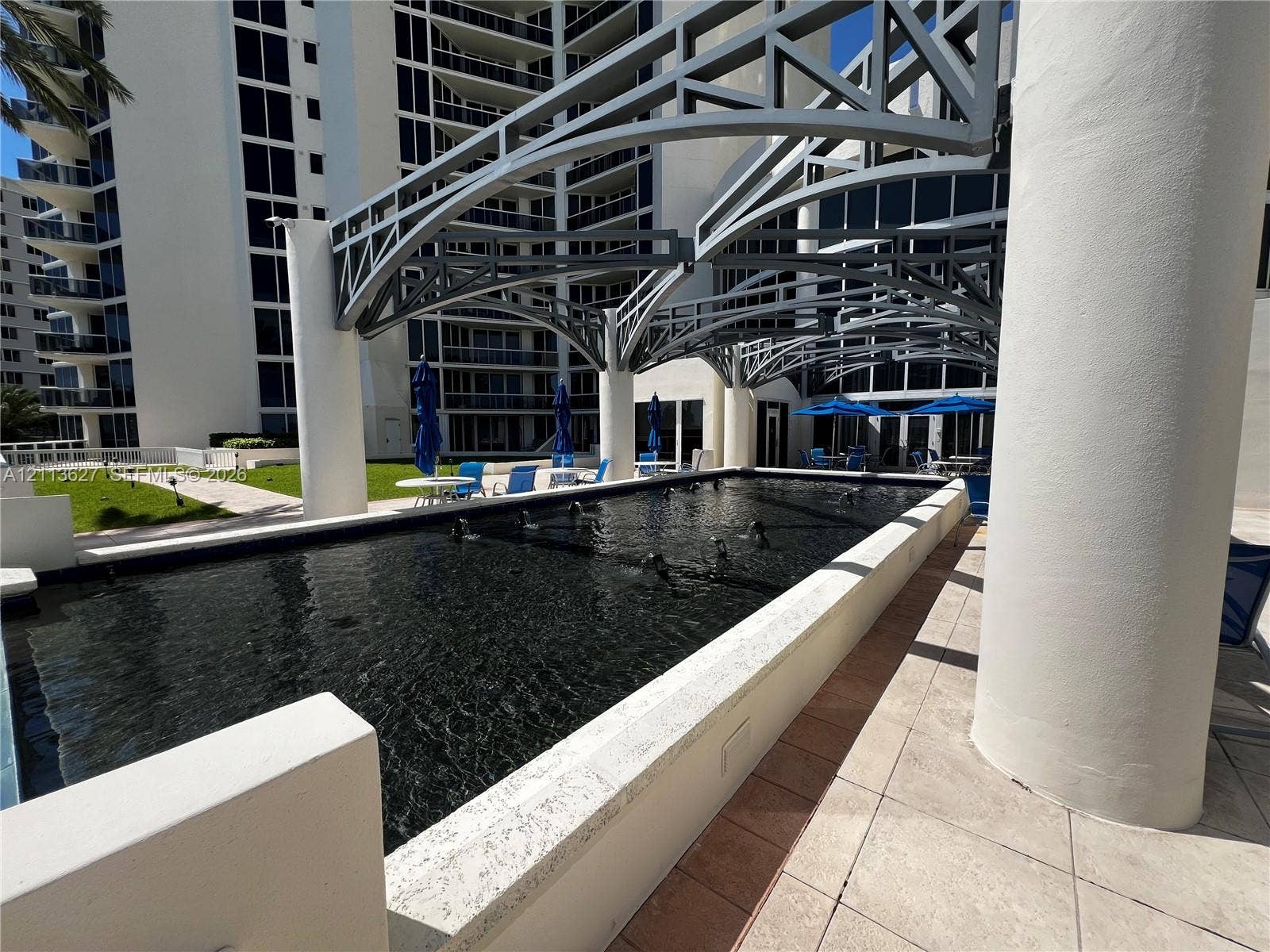 Rectangular reflective pool with built-in water features, architectural overhead metal framing, cylindrical support columns, and light-toned stone tile paving