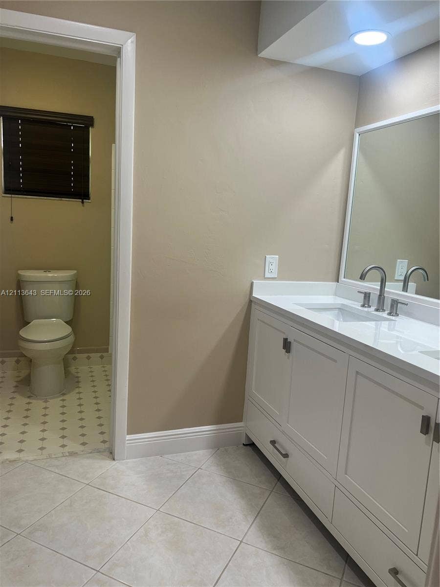 White vanity with a rectangular sink, stone-finish countertop, dual-handle faucet, large wall-mounted mirror, and tile flooring