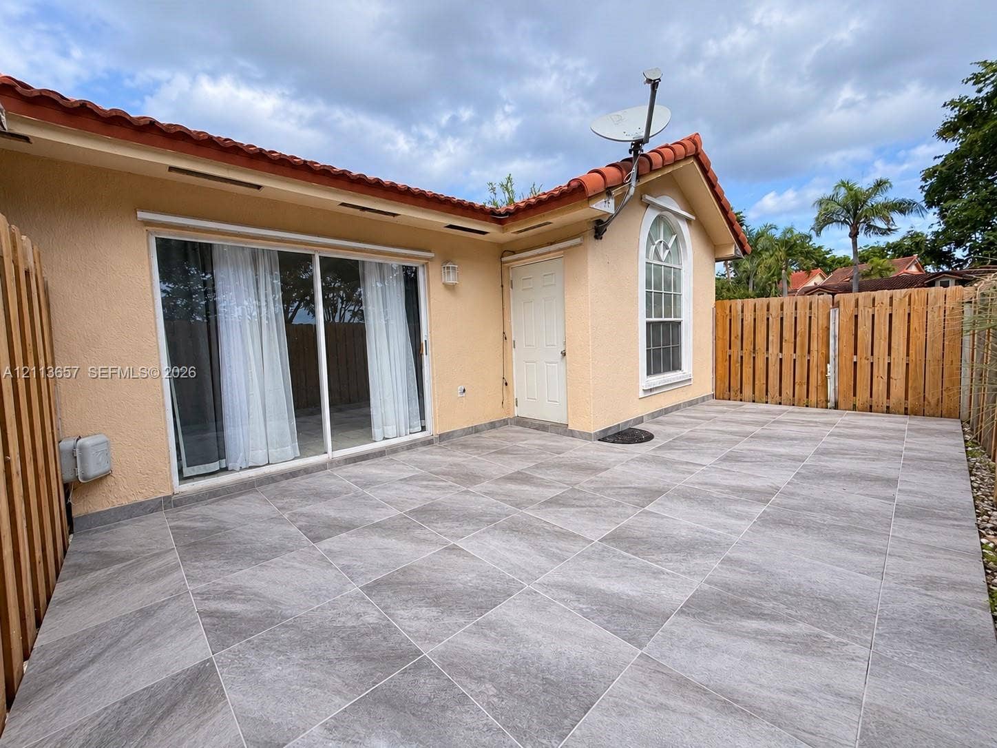 Stucco exterior with a tiled roof, expansive gray-tiled patio, sliding glass door, and wooden privacy fencing