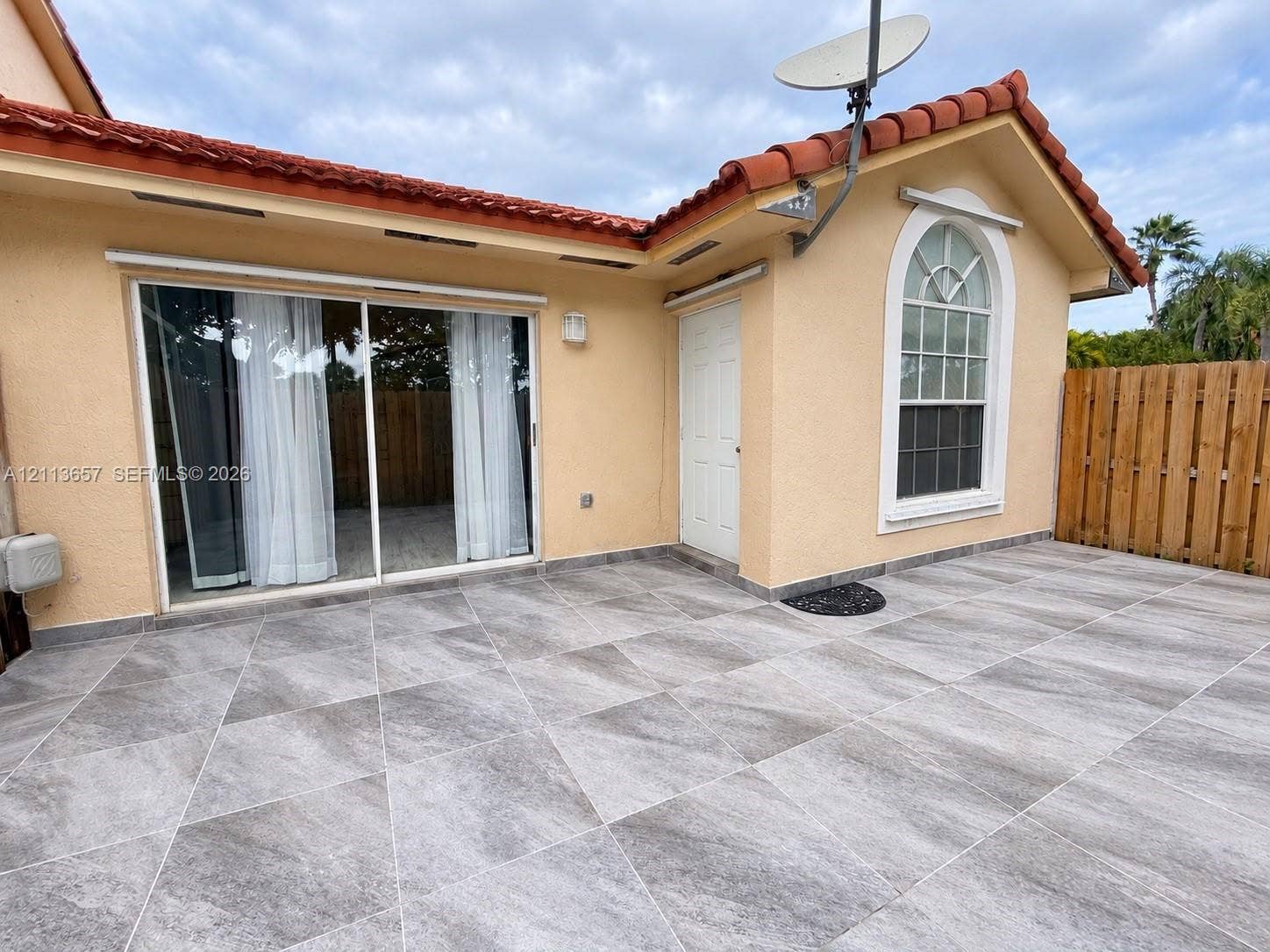Stucco exterior featuring red clay roof tiles, large-format grey tile patio, sliding glass door, arched window, and wood privacy fencing