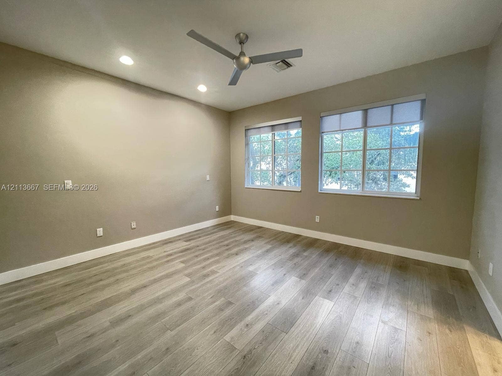 Neutral-toned interior featuring wood-finish flooring, dual windows with multi-pane glazing, and a ceiling-mounted brushed metal fan with recessed lighting