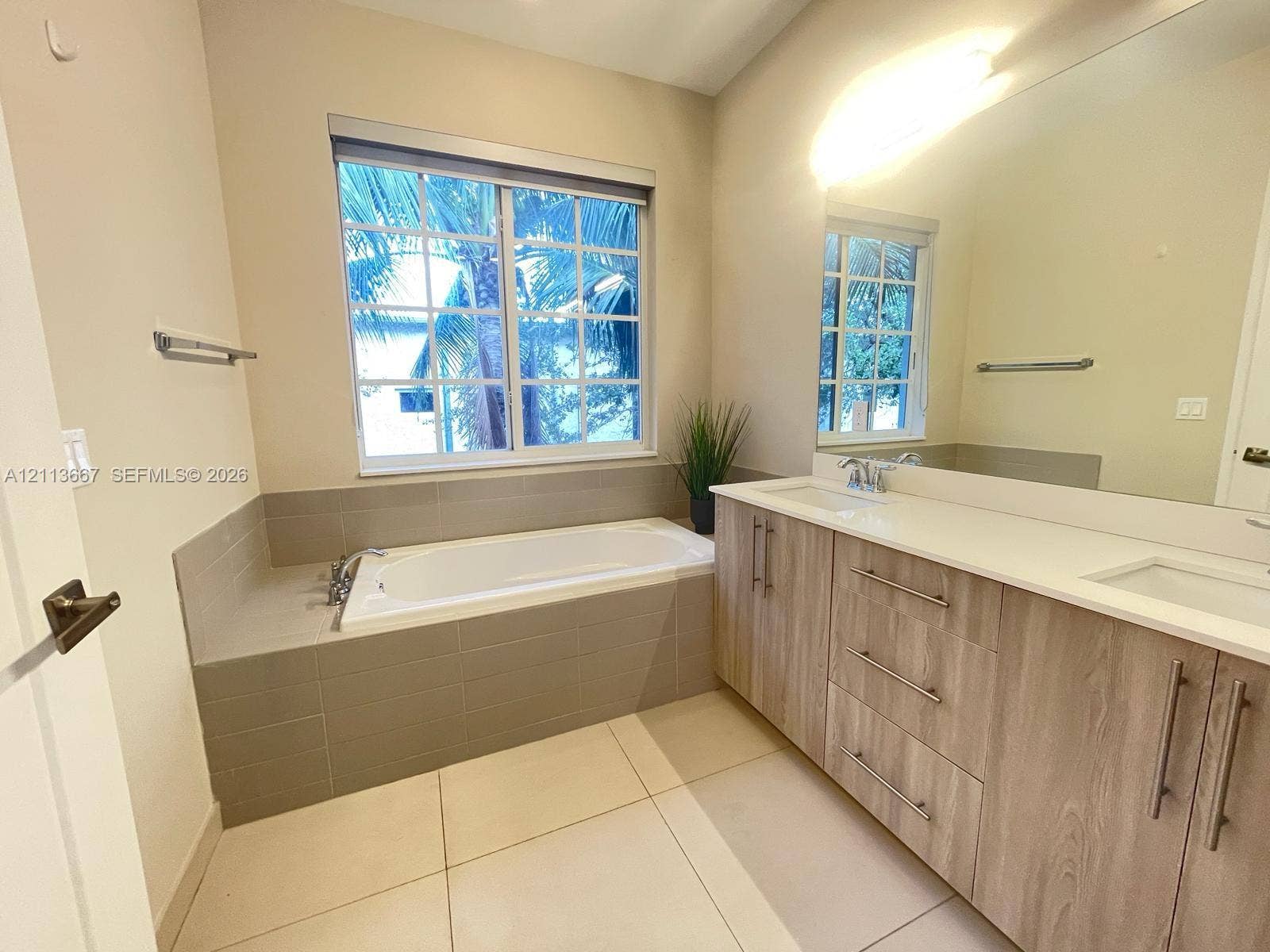 Bathroom featuring a dual-sink vanity with wood-finish cabinetry, a drop-in soaking tub with a tiled surround, a large wall-mounted mirror, and tile flooring