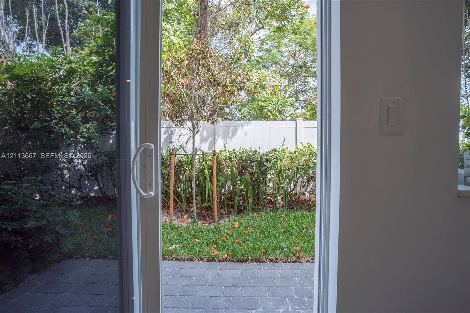 Sliding glass door opening to a paved patio, bordered by a lawn, mature shrubs, and a privacy fence