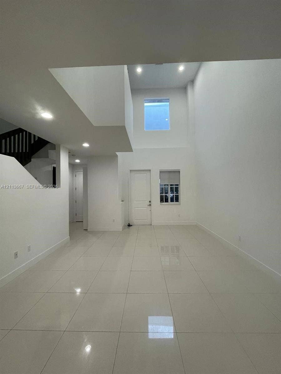 High-ceiling interior featuring glossy tile flooring, recessed lighting, and a staircase with a dark wood-finish railing