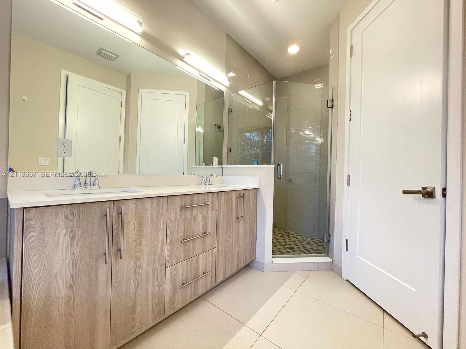 Dual-vanity cabinetry with wood-finish doors, solid-surface countertops, full-width mirror, and a glass-enclosed shower with tiled flooring